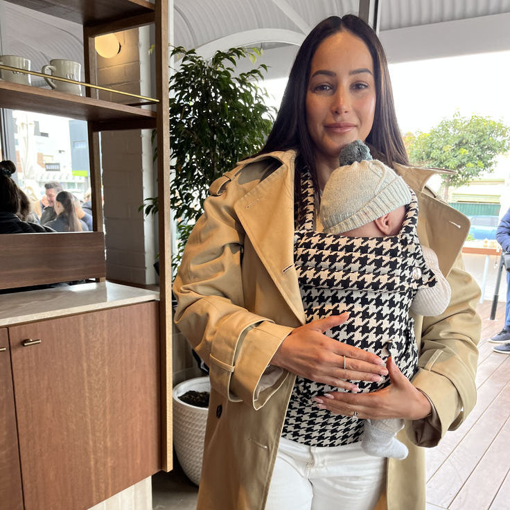Woman in a beige trench coat holding a baby in a sling, standing indoors.