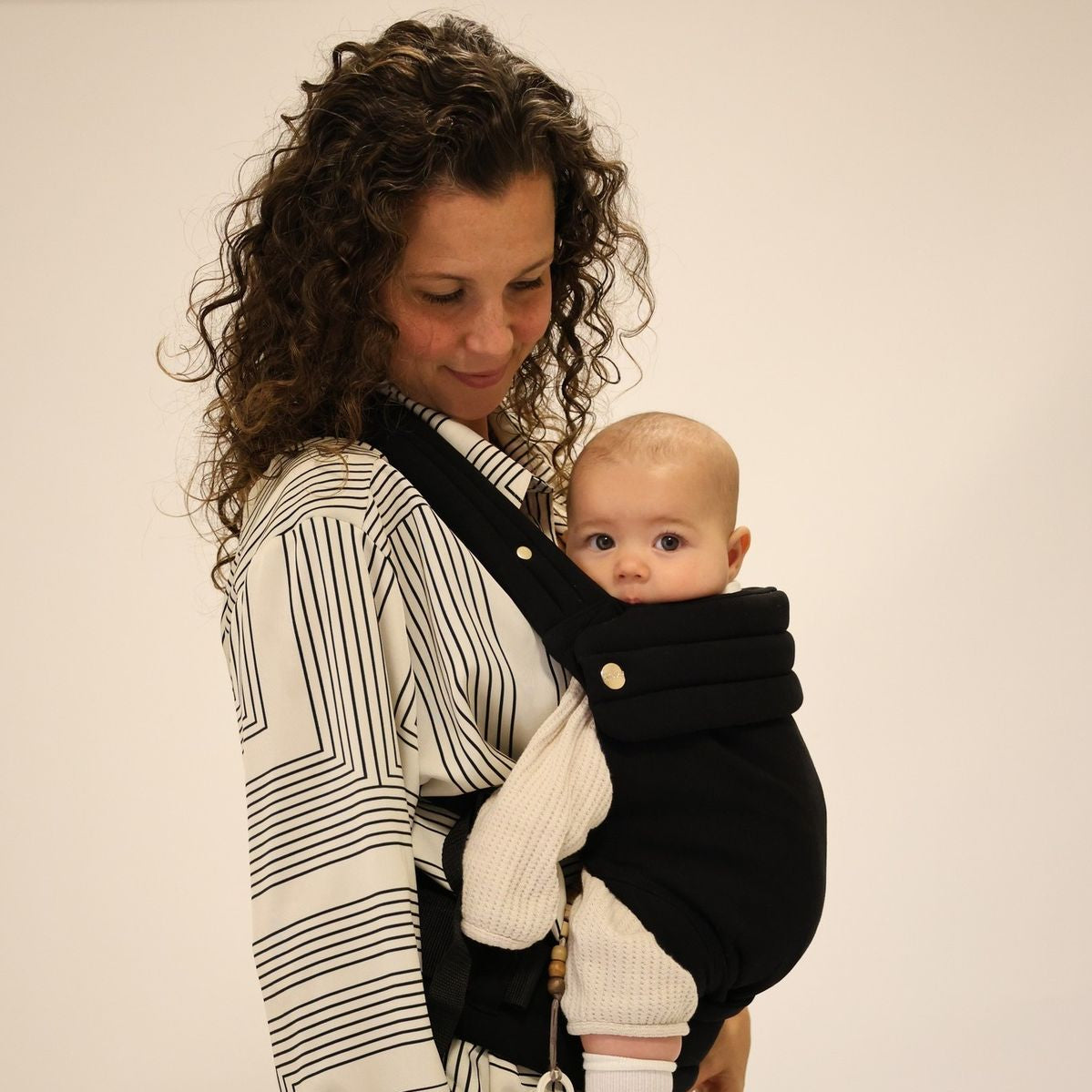 Woman holding a baby in a black baby carrier against a plain background
